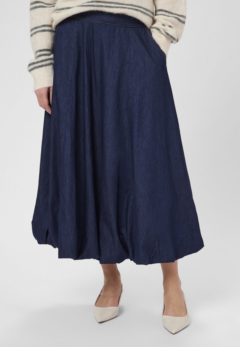 Fustă midi din denim de culoare albastru închis, cu pliseuri subtile și buzunare laterale. Asortată cu balerini de culoare deschisă și un pulover în dungi.
