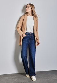 Abrigo de mezcla de lana beis sobre un suéter blanco, combinado con jeans de campana azules. La modelo lleva zapatillas blancas. Texturas suaves y diseño casual.