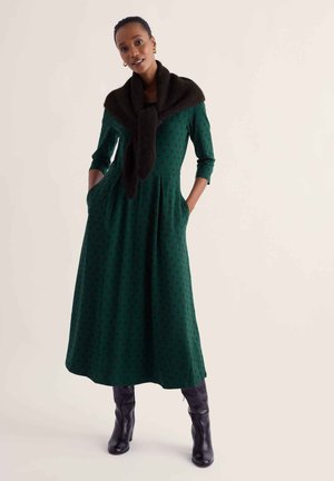 Femme debout les mains dans les poches, portant une longue robe verte à pois noirs, des bottes noires et une écharpe noire sur les épaules.
