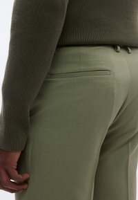 Grønne bukser med en glat tekstur, der har en baglomme og en skræddersyet pasform. Bæres med en mørkegrøn, ribstrikket sweater.