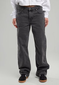 Tummanharmaat leveät farkut korkealla vyötäröllä, viisi taskua ja hienovarainen haalistuminen. Kangas näyttää rakenteelliselta ja kestävältä. Päällä mustat kengät.