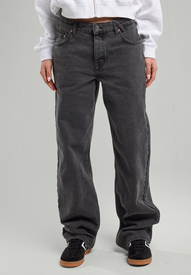 Tummanharmaat leveät farkut korkealla vyötäröllä, viisi taskua ja hienovarainen haalistuminen. Kangas näyttää rakenteelliselta ja kestävältä. Päällä mustat kengät.