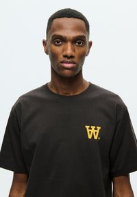 Black cotton t-shirt with short sleeves, featuring a small yellow logo on the left chest. Smooth texture, standard crew neck design.