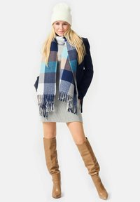 Woman wearing white knit hat, navy coat, gray sweater dress, blue and beige checkered scarf, and tan knee-high boots standing on white background.