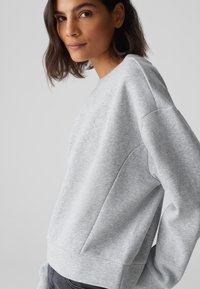 Grauer Hoodie aus weichem Stoff, mit einem gerippten Ausschnitt und einem kleinen Herz-Logo auf der Brust. Angenehmer, lässiger Schnitt mit seitlichen Details.