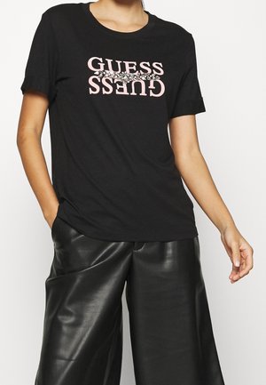 Black short-sleeve t-shirt featuring "GUESS" logo in pink, accented with rhinestones. Paired with loose-fitting black leather pants.
