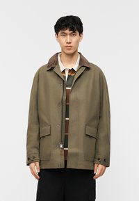 Νέος άνδρας που φοράει χακί πράσινο μπουφάν με καφέ γιακά από κορντόρι πάνω από μια ριγέ πόλο μπλούζα, στέκεται μπροστά σε απλό λευκό φόντο.