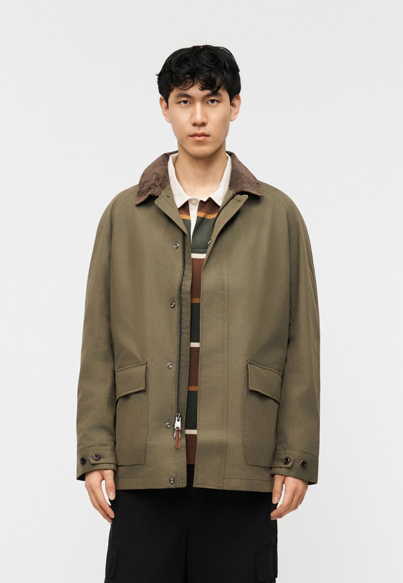 Νέος άνδρας που φοράει χακί πράσινο μπουφάν με καφέ γιακά από κορντόρι πάνω από μια ριγέ πόλο μπλούζα, στέκεται μπροστά σε απλό λευκό φόντο.