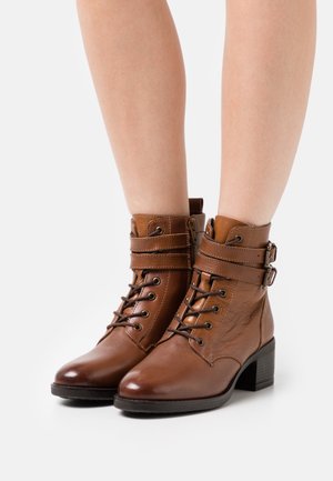 Braune Lederstiefeletten mit Schnürung, geschnallten Riemen und einer strukturierten Oberfläche. Der Blockabsatz und die abgerundete Zehenpartie vervollständigen das Design.