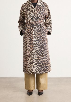 Abrigo tipo trench con estampado de leopardo y cinturón, llevado sobre pantalones beige de pierna ancha y tacones negros de punta afilada, con las manos en los bolsillos del abrigo.