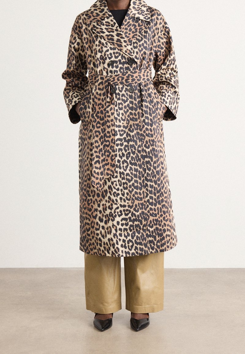 Trench-coat ceinturé à motif léopard porté avec un pantalon large beige et des escarpins noirs à bouts pointus, mains dans les poches du manteau.