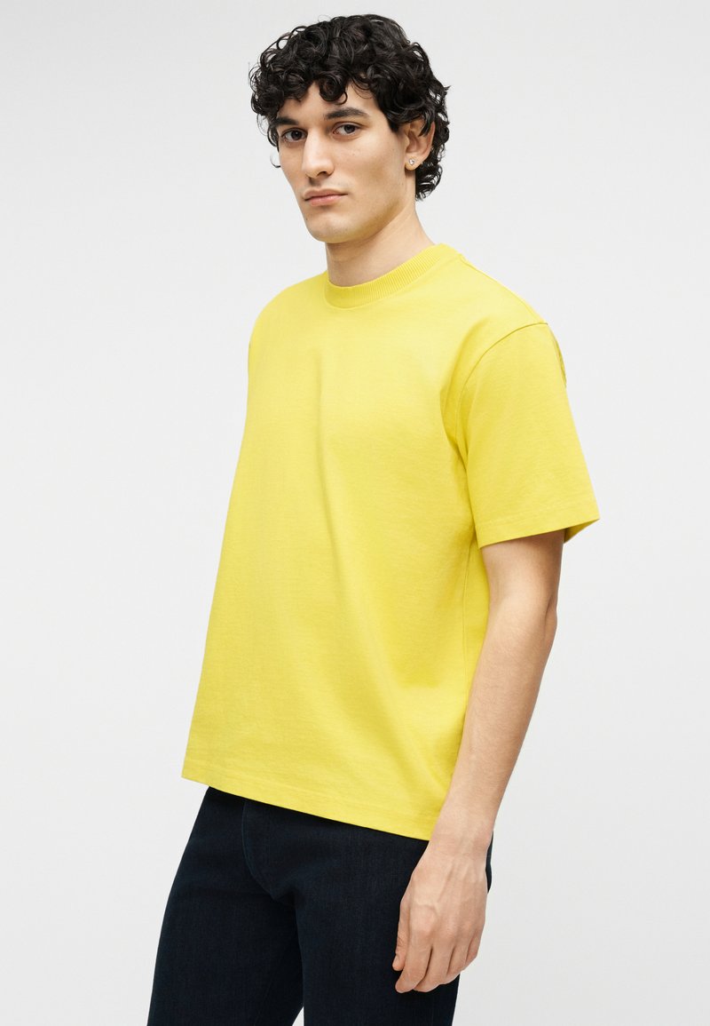 Giovane con capelli ricci che indossa una maglietta gialla a maniche corte semplice e pantaloni scuri, in piedi davanti a uno sfondo bianco.