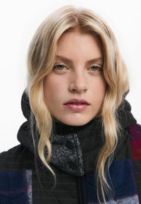 Giaccone puff nero con colletto grigio a fantasia, caratterizzato da grandi bottoni e accenti di patchwork colorati. Lunghi capelli biondi e mossi visibili.