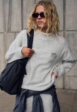 Mujer rubia con gafas de sol negras, suéter gris con texto "self athleisure", llevando un bolso de cuadros, con camisa de cuadros atada alrededor de la cintura.