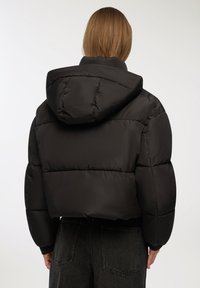 Czarna kurtka ocieplana z kapturem, o pudełkowatym kształcie, z elastycznymi mankietami i gładką, błyszczącą teksturą. Widok z tyłu ukazuje pikowany wzór.
