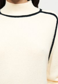 Κρεμ πλεκτό πουλόβερ με ψηλό γιακά, μαύρο περιθώριο κατά μήκος του λαιμού και των ραφών στους ώμους, μαλακή υφή και χαλαρή εφαρμογή.