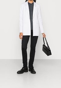 Blazer bianco con rever a punta su una camicia scura con bottoni, abbinato a pantaloni neri slim e scarpe nere, mentre tiene una borsa nera.