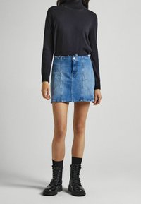 Mini jupe en denim bleu clair au design ajusté, avec un ourlet brut et une fermeture à boutons sur le devant ; associée à un top noir à manches longues et des bottes noires.