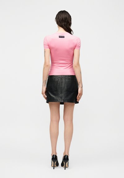 Γυναίκα που στέκεται με την πλάτη προς τα εμπρός φορώντας ροζ εφαρμοστή κοντομάνικη μπλούζα με ετικέτα, μαύρη δερμάτινη φούστα και μαύρες ψηλοτάκουνες γόβες.