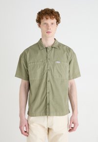 Chemise à manches courtes en vert olive, en coton, avec une fermeture à boutons, deux poches poitrine et une étiquette sur la poche gauche.