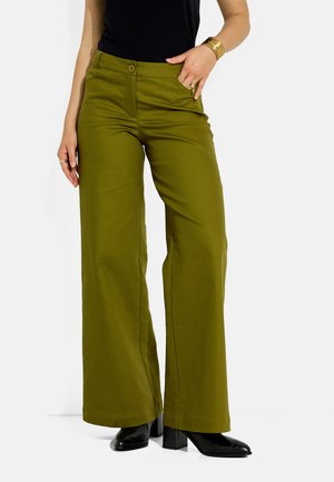 Mujer vistiendo pantalones anchos de color verde oliva, botas negras con tacón, una camiseta negra y pulseras doradas sobre un fondo blanco.