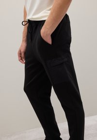 Schwarze Jogginghosen mit elastischem Bund, Kordelzug, seitlichen Taschen und einer strukturierten aufgesetzten Tasche am Oberschenkel. Glatte Stoffoberfläche.
