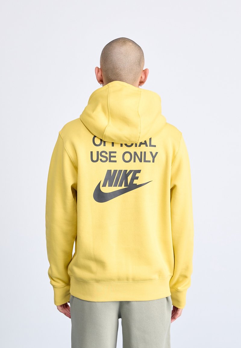 Žlutá mikina s kapucí z měkké tkaniny, na zadní straně velký nápis „POUZE PRO OFICIÁLNÍ POUŽITÍ“ a černé logo Nike.