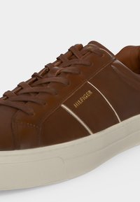 Кафяв кожен спортен обувка с връзки, бяла подметка и златно лого "HILFIGER" отстрани.