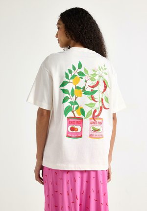 Kvinde iført hvid t-shirt med citron- og chiliplanter, der vokser ud af dåser mærket "Always Fresh", kombineret med en lyserød nederdel med chiliprint.