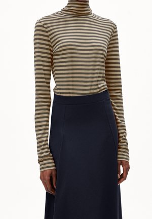 Personne portant un col roulé à rayures horizontales beige et grises et une jupe taille haute bleu marine foncé, debout les bras détendus sur les côtés.