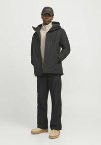 Chaqueta negra impermeable con capucha y bolsillos con cremallera, combinada con pantalones cargo negros y botas de trabajo color marrón. Tejido texturizado y diseño casual.