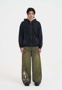 Zwarte rits hoodie met een zachte textuur en logodetail aan de zijkant, gecombineerd met losse olijfgroene jeans met een grafisch ontwerp.