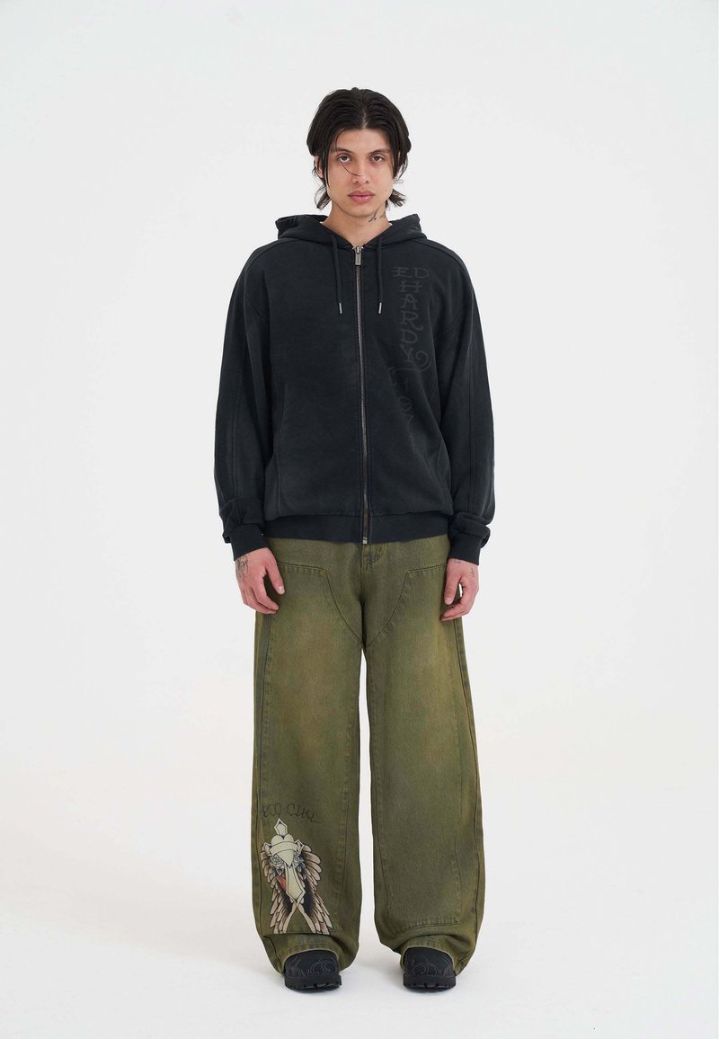 Schwarze Zip-Hoodie mit weicher Textur und Logodetail an der Seite, kombiniert mit lässigen olivgrünen Jeans mit grafischem Design.