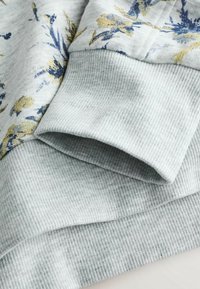 Tissu gris clair avec texture côtelée sur les poignets et l'ourlet, présentant un imprimé floral aux tons bleus et jaunes sur un tissu lisse.