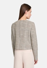 Cropped jacket in textured beige fabric with a woven pattern. Features long sleeves and a square neckline. Smooth finish and minimalist design.