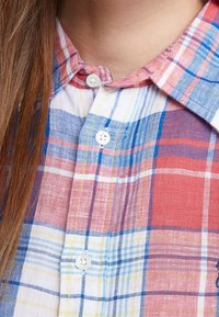 Camicia a scacchi con colori rosso, blu e giallo, con design a bottoni e tessuto leggermente strutturato. Collo con bottoni visibili.