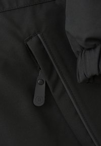 Detalj av svart tygjacka med en dragkedjeficka och en gummihandtag, som visar en slät textur och rena linjer.