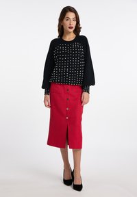 Zwarte gebreide trui met opstaande mouwen en witte kraalaccenten; rode midi rok met een split aan de voorkant en knoopdetails; zwarte hakken.