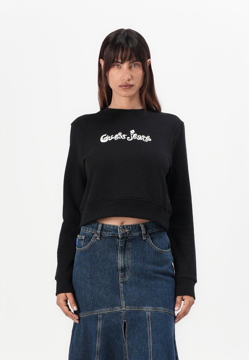 Sweatshirt preto cropped com gráfico branco "Guess Jeans". Combinado com uma saia de ganga com fenda frontal e detalhes de costura visíveis.