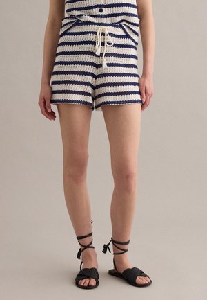 Mujer con pantalones cortos de punto a rayas azul marino y blanco con cordón y sandalias planas negras con cordones, de pie frente a un fondo liso.
