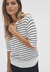 Lagan, bijeli pulover na izvlačenje s tamnoplavim prugama, ima opušteni kroj, kratke rukave i rebrasti rub. Opremljen zlatnim narukvicom.