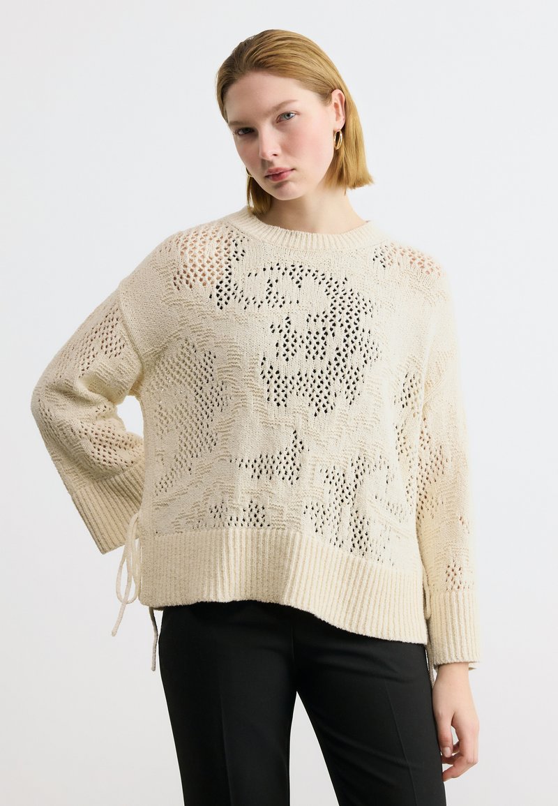 Νεαρή γυναίκα με κοντά ξανθά μαλλιά φορώντας εκρού πλεκτό πουλόβερ με λουλουδένιο σχέδιο και μαύρο παντελόνι, στέκεται με το ένα χέρι στη μέση.