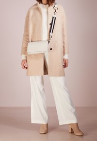 Cappotto beige texturizzato con taglio dritto, indossato su un maglione bianco e pantaloni a gamba larga. Presenta una borsa a tracolla bianco e nero.