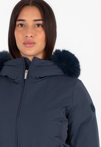 Giacca imbottita blu navy con cappuccio in pelliccia sintetica, dotata di zip frontale e design strutturato. Texture liscia e silhouette arrotondata.