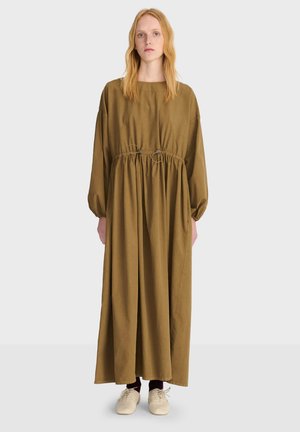 Donna con lunghi capelli biondi che indossa un abito lungo e morbido verde oliva con maniche lunghe e scarpe beige chiare, in piedi su sfondo bianco.