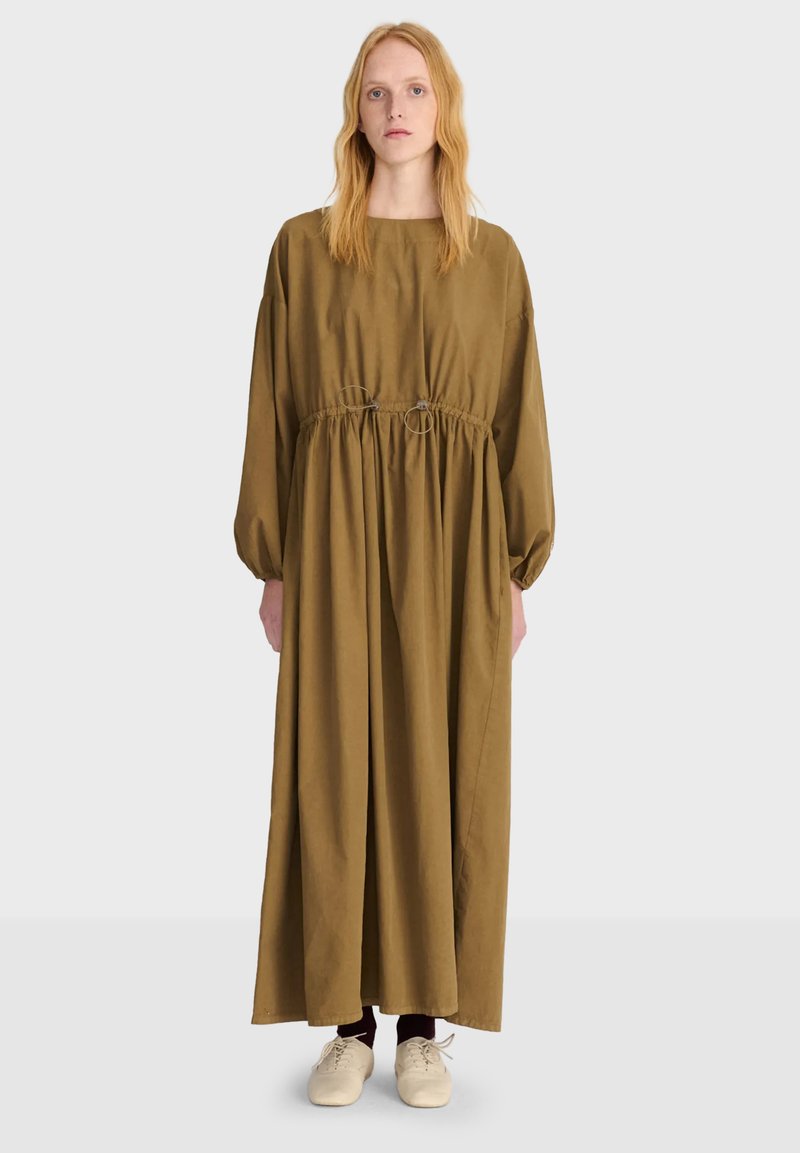 Femme aux longs cheveux blonds portant une robe maxi ample à manches longues de couleur vert olive et des chaussures beige clair, debout devant un fond blanc.
