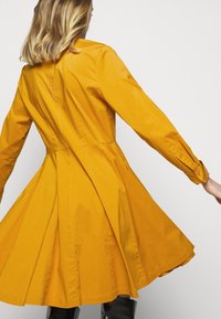 Żółta sukienka z gładkiego materiału, z rozkloszowanym dołem, dopasowaną górą, długimi rękawami i guzikami przy mankietach.