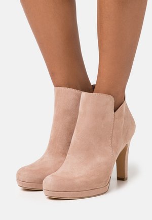 Une paire de bottines à talons hauts en daim rose clair portées aux pieds, avec un bout arrondi et des talons blocs fins.