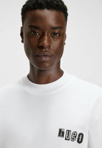 Young man with short curly hair wearing a white sweatshirt with "hugo" printed in black mixed fonts on the chest.