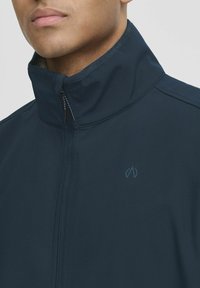 Marine modra zip-up jakna z visokim ovratnikom, ki ima subtilen logotip na prsih in gladko, sleek teksturo tkanine.